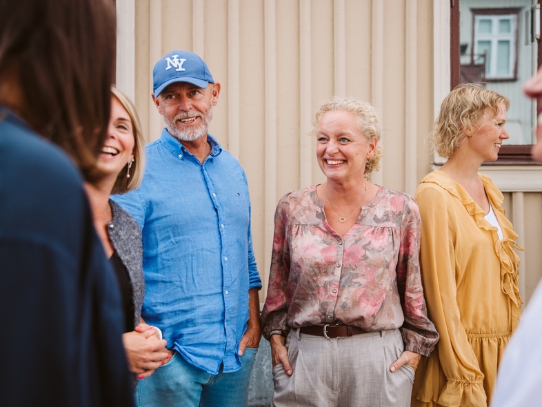 En grupp vuxna står utomhus framför en beige vägg och samtalar.