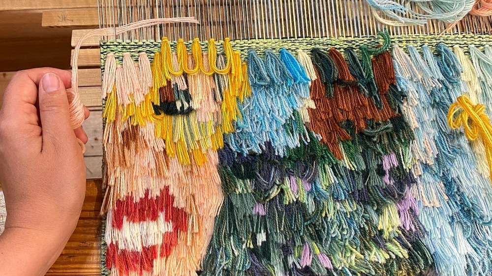 En hand väver färgglada garntrådar på en vävstol, med mönster i gult, blått, rött och grönt.