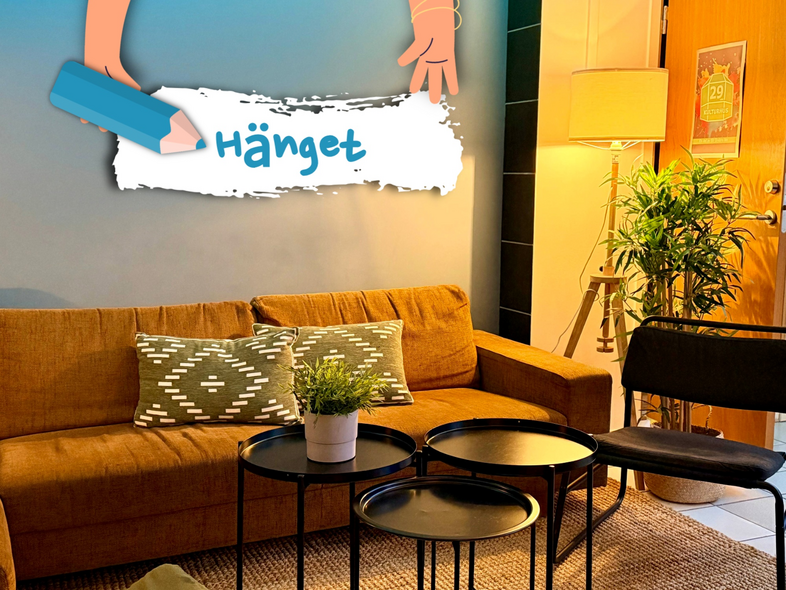 Mysigt vardagsrum med brun soffa, gröna kuddar, svarta bord och växt. Texten 'Hänget' på väggen.