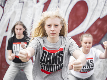 Tre unga personer dansar energiskt framför en graffitivägg, med fokus på en tjej i grå t-shirt.