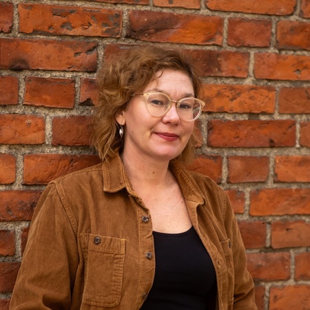 Kvinna med glasögon och brunt hår står mot en tegelvägg, klädd i brun jacka och jeans.
