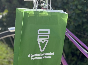 En grön papperspåse med Studieförbundet Vuxenskolans logotyp på hänger på styret till en rosalila cykel som står på en solig cykelväg bredvid en häck.