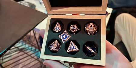 En hand håller en öppen trälåda med sex olika tärningar, troligen för rollspel, i en vadderad insats.