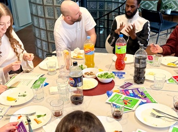 En grupp unga vuxna sitter runt ett bord och spelar ett kortspel, med mat och dryck på bordet.