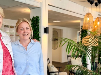 Två personer står tillsammans i ett modernt, ljust kontorslandskap med gröna växter och soffor.