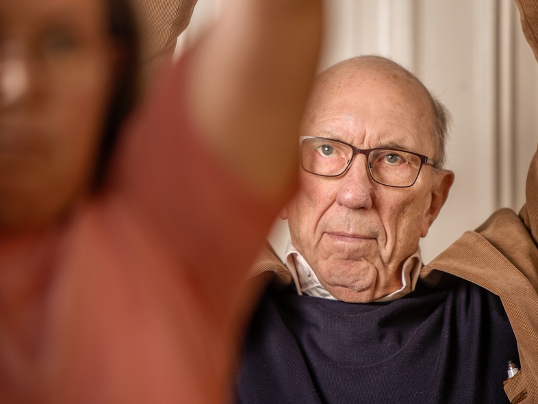 Äldre man med glasögon sträcker armarna över huvudet, suddig person i förgrunden.