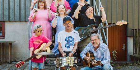 En grupp människor poserar utomhus framför ett hus, flera håller musikinstrument.