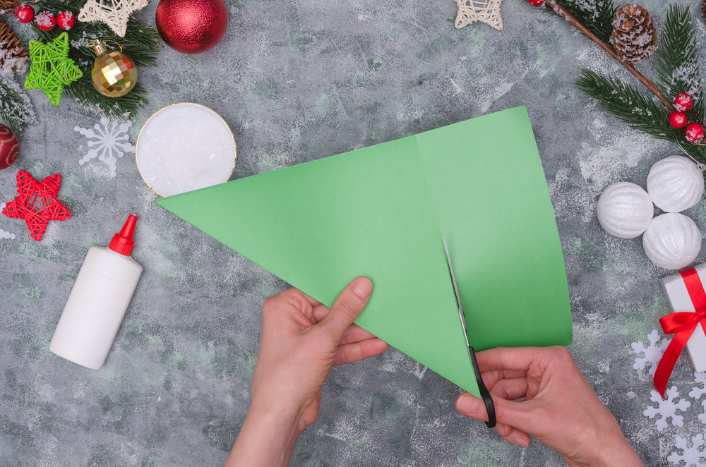 Två händer klipper en grön pappersbit i form av en triangel, omgiven av juldekorationer och lim.
