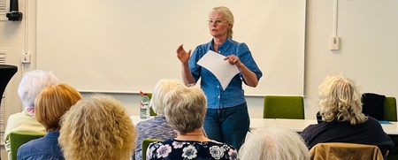 En kvinna håller en presentation framför en grupp äldre personer i ett mötesrum med gröna stolar.