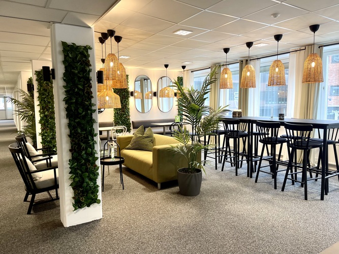 Modern loungeyta med gröna växter, soffor, höga bord och stolar samt stora fönster med naturligt ljus.