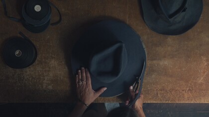 En person klipper en mörk hatt på ett träbord, sedd uppifrån. Bandrullar ligger bredvid.