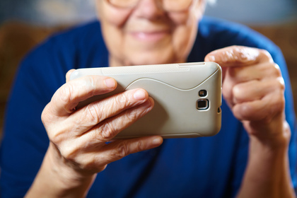 En äldre person håller och använder en smartphone med båda händerna, leende syns i bakgrunden.