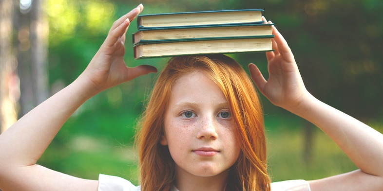Flicka i vit klänning balanserar tre böcker på huvudet utomhus i en grön skogsmiljö.