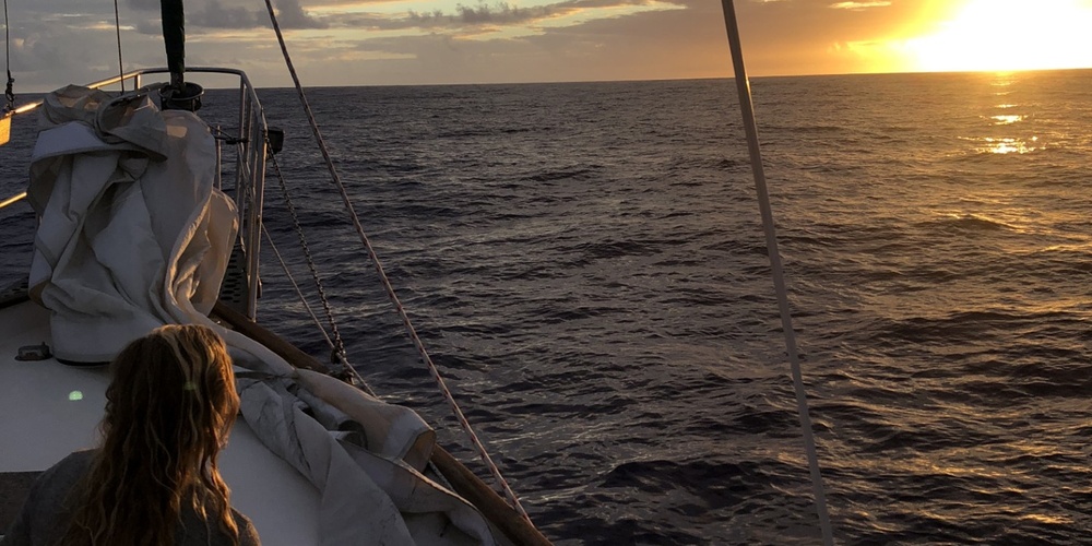 En person sitter på en segelbåt och tittar ut över havet mot en solnedgång.