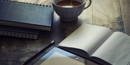 Ett skrivbord med en kopp kaffe, anteckningsböcker, en penna och en surfplatta.