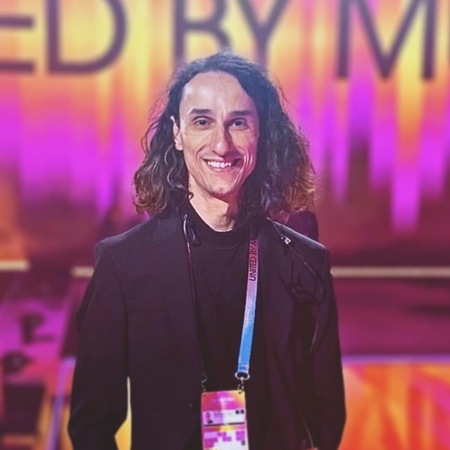 En person med långt lockigt hår och svart kavaj står leende framför en färgglad bakgrund med text.