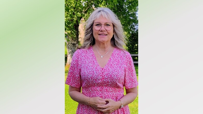 Kvinna med blont hår i rosa blommig klänning står utomhus på gräsmatta med träd i bakgrunden.