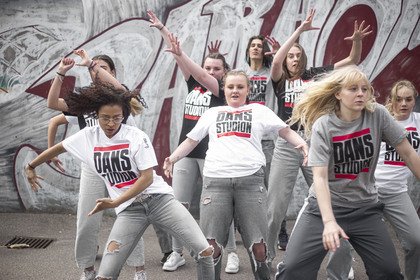 En grupp unga kvinnor dansar energiskt utomhus framför en graffitivägg, alla bär t-shirts med texten 'DANS STUDIO'.