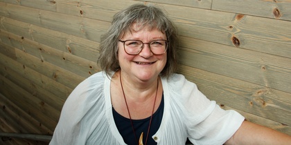 Leende kvinna med glasögon och grått hår står mot en trävägg.