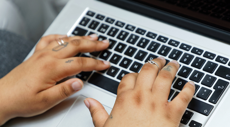 Två händer med ringar och tatueringar skriver på ett svartvitt tangentbord på en bärbar dator.