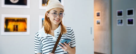 Ung kvinna med glasögon och mössa står leende i ett konstgalleri och håller en mobiltelefon.