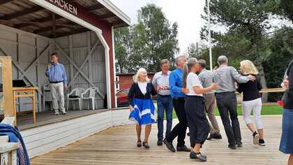 Folk dansar på en träscen utomhus medan en man talar i mikrofon på en upphöjd scen.