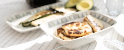 Ett dukat bord med grillad majs, sparris, kyckling, avokado och en vas med lila blommor i solljus.