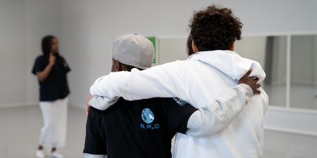 Två personer står med armarna om varandra i ett ljust rum, en tredje person syns i bakgrunden.