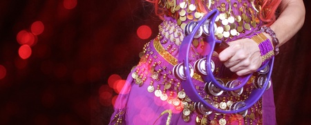 Person i lila magdansdräkt med guldmynt och tamburin, mot röd bakgrund med ljuseffekter.