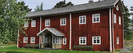 Fågelsjö gammelgård bortom Åa en röd Hälsingegård med vita fönster och veranda, omgiven av gräs och träd under en ljus himmel.