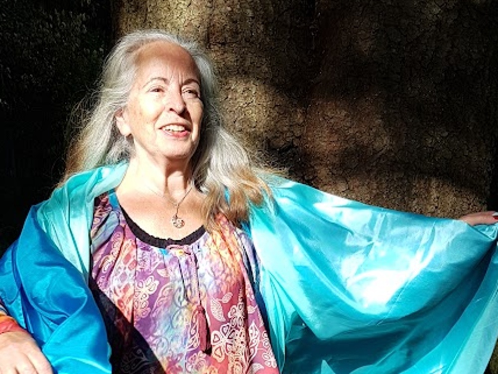 Cirkelledare Elisabeth Hjulström med långt grått hår står barfota utomhus, bär färgglad tunika och blå sjal.