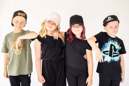 Fyra barn i kepsar poserar tillsammans mot en vit bakgrund, två pojkar och två flickor.