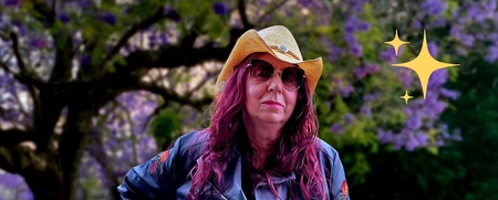 Person med cowboyhatt, solglasögon och svart skinjacka med silverdetaler och röda blommor står utomhus framför grönskande träd och tittar in i kameran. Personen har långt rött vågigt hår. 