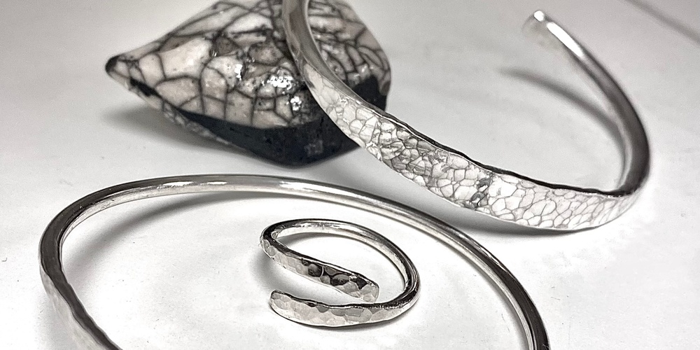 Tre silversmycken, två armband och en ring, ligger på ett vitt underlag med en svartvit sten i bakgrunden.