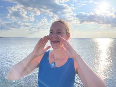 Person i blå linne står vid en sjö med händerna upp, solnedgång och molnig himmel i bakgrunden.