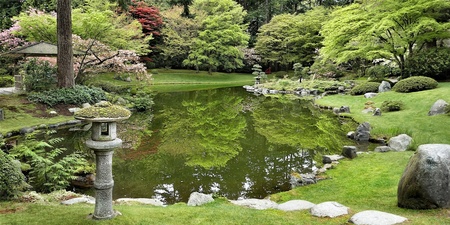 En lugn japansk trädgård med damm, gröna träd, stenar och en stenlykta vid vattnet.