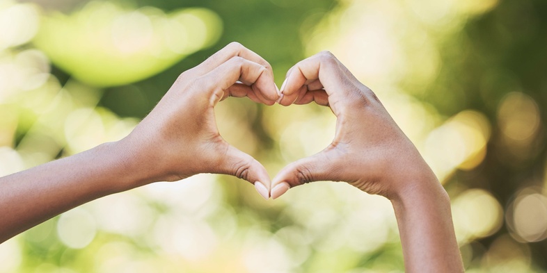 Två händer formar ett hjärta mot en suddig, grön bakgrund utomhus.