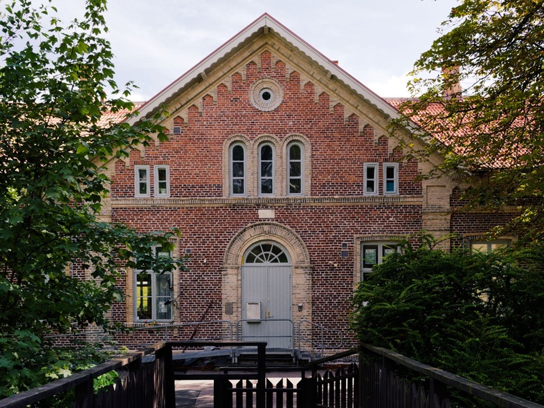 En gammal tegelbyggnad med spetsigt tak, sedd från en träbro omgiven av grönska.