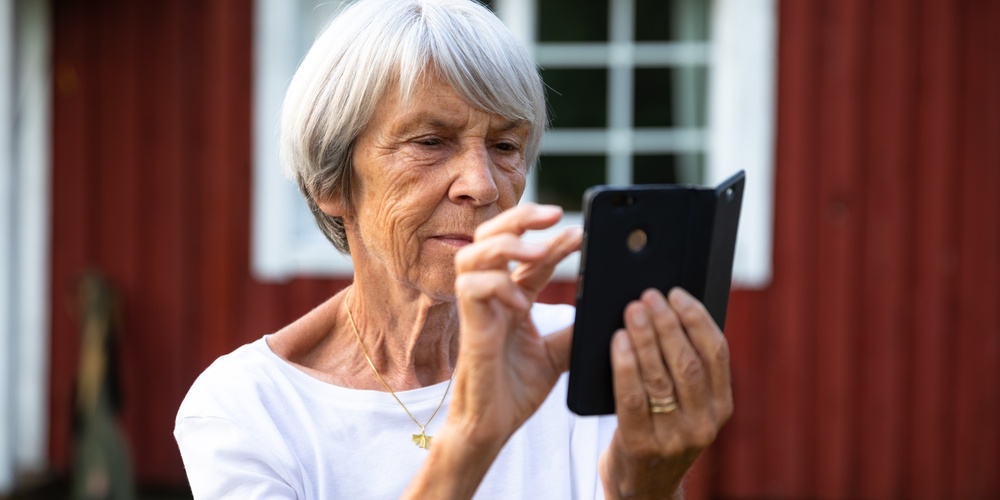 Äldre kvinna med grått hår använder en smartphone utomhus framför ett rött trähus.