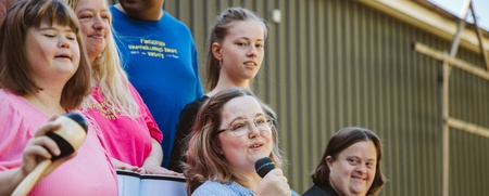 En grupp människor, flera med mikrofon och musikinstrument, står utomhus och sjunger tillsammans.