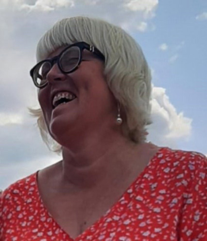 Leende kvinna med ljust hår och glasögon i röd mönstrad klänning utomhus mot blå himmel.
