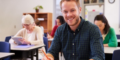 Leende man sitter vid skolbänk i klassrum, skriver i anteckningsbok. Andra vuxna studenter i bakgrunden.