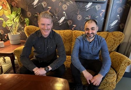 Två män sitter på en gul soffa i ett rum med mönstrad tapet och växter.