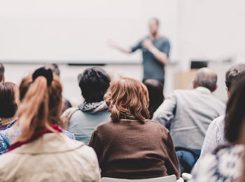 En grupp människor sitter och lyssnar på en person som håller en presentation i ett ljust rum.