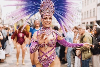 Kvinna i färgglad karnevalsdräkt med fjädrar dansar på gata inför publik under en parad.