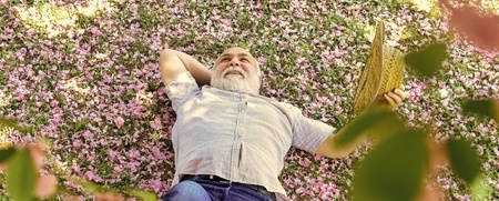 En äldre man ligger avslappnat på marken täckt av rosa blomblad och håller en bok i handen.