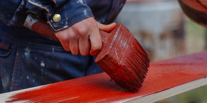 En person målar en träplanka röd med en bred pensel, iförd blå arbetskläder.
