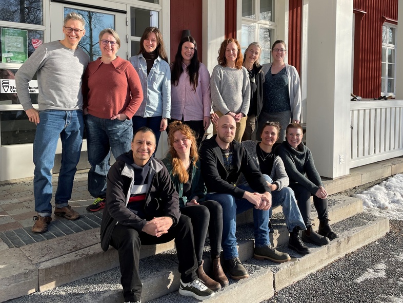 En grupp på tolv personer poserar utomhus framför en röd byggnad i vintersol.