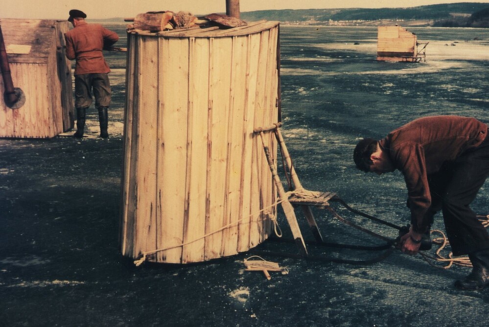 Två personer arbetar vid små träkojor på en frusen sjö, troligen för isfiske. Vintrig miljö med is och snö.