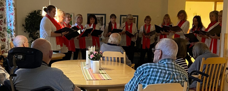 En kör sjunger för äldre personer som sitter runt ett bord på ett äldreboende.